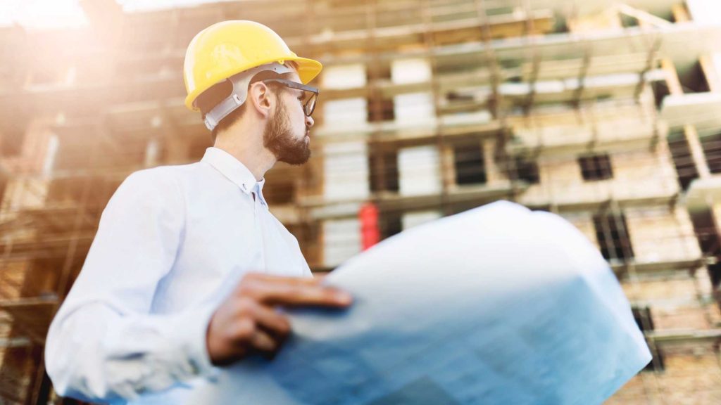 Man wearing a hardhat, holding a building plan in front of a building with scaffolding