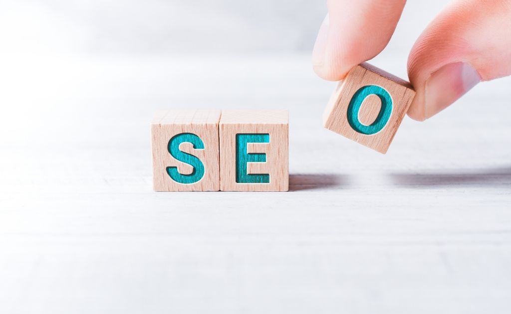 3 wooden building blocks spelling out SEO on them in blue. Hand placing O beside E. Local SEO helps to create a business presence.