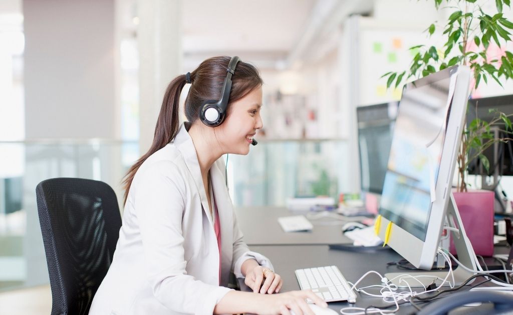 Smiling woman sitting at a computer screen, answering business calls using a headset.