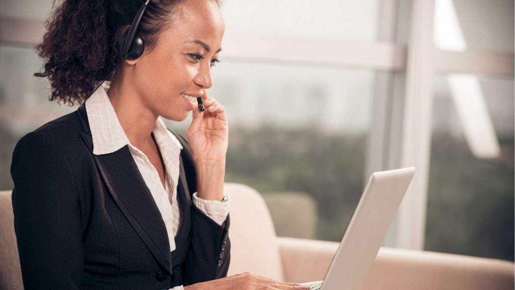 Smiling woman working as a UK virtual receptionist, chatting to a customer on a headset, sitting in front of a laptop.