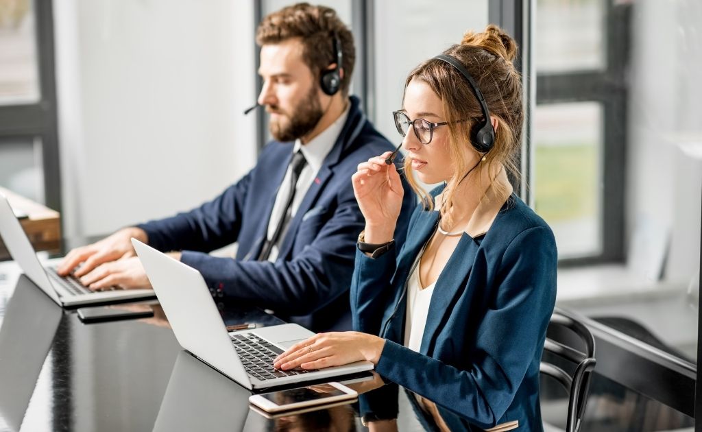 A woman and a man, both sitting in front of a laptop, answering calls using a headset.