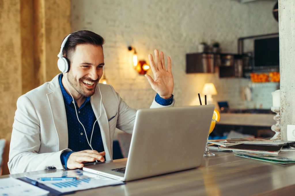 Smiling man with white headphones sat working at home on laptop waving to laptop camera