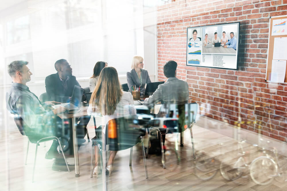 Team sat at desk on video conference call with others on TV