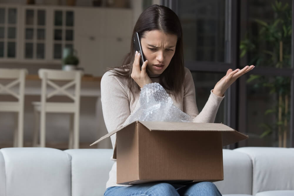 Woman sitting on sofa on phone looking angry check received box, damaged or broken goods in parcel, talks to customer support