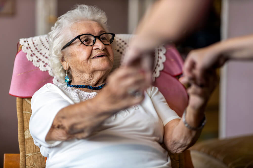 An older woman sits in a chair, holding her hand lovingly.