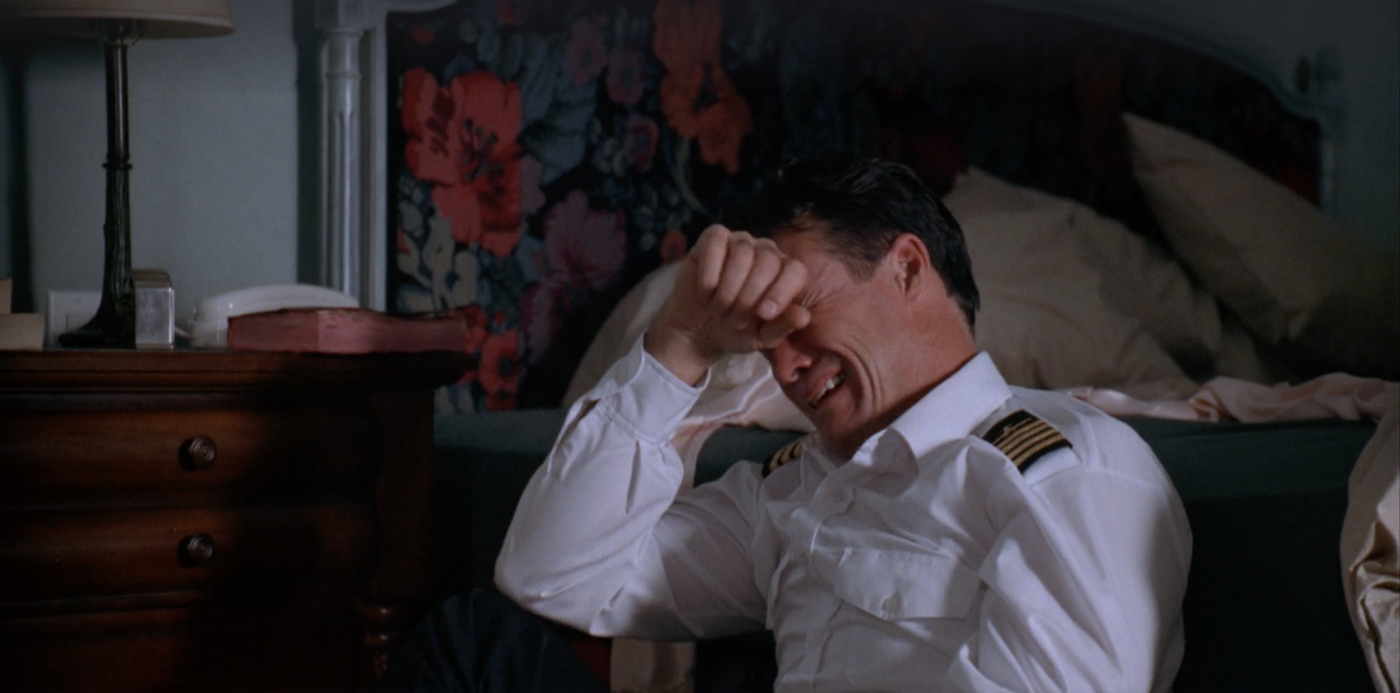 Rayford in a pilot's uniform leaning against a bed, holding his right hand to his eyes, and crying.