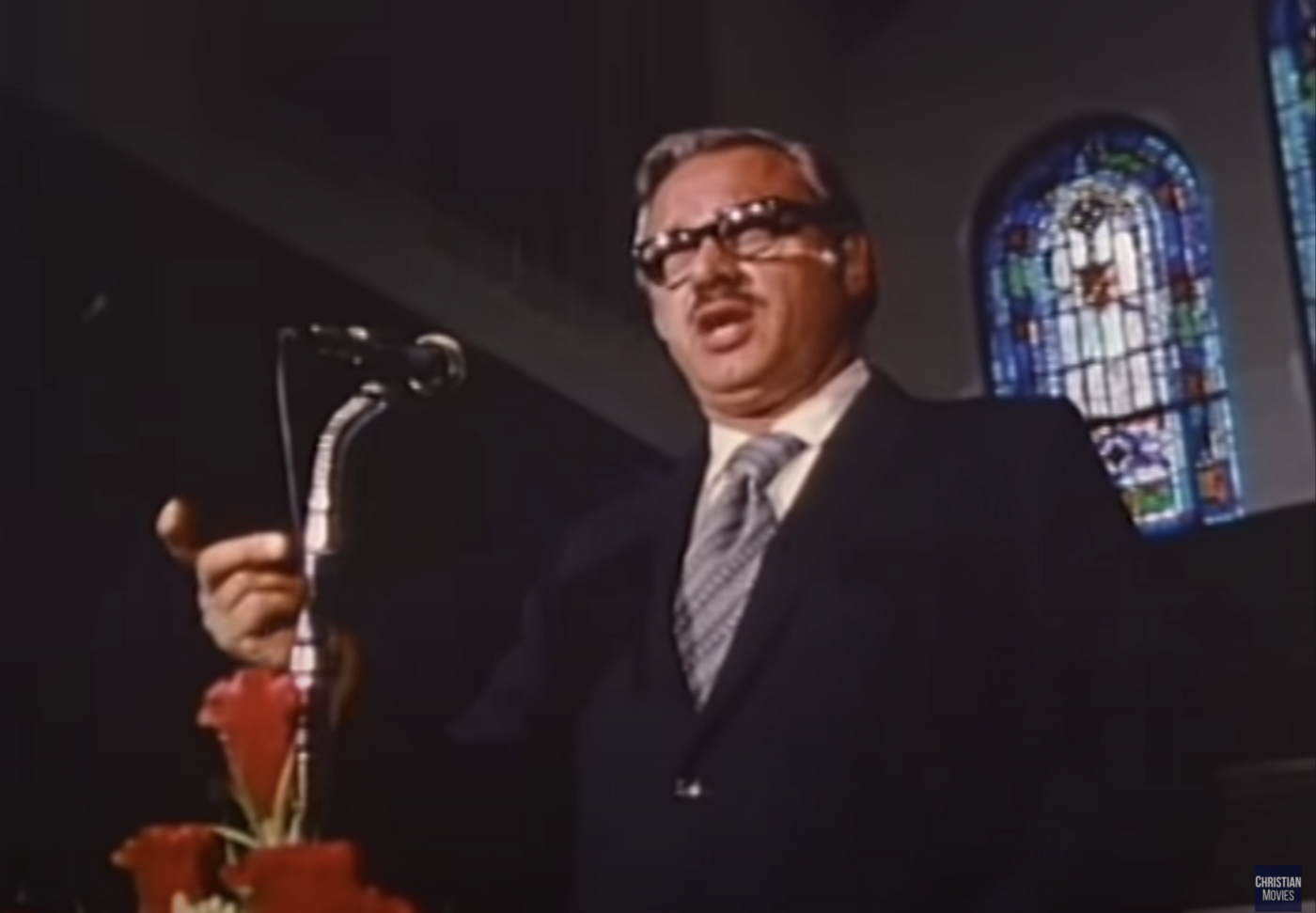 Reverend Turner, a grey-haired man in a suit and thick glasses with a thin moustache, preaching at a pulpit.