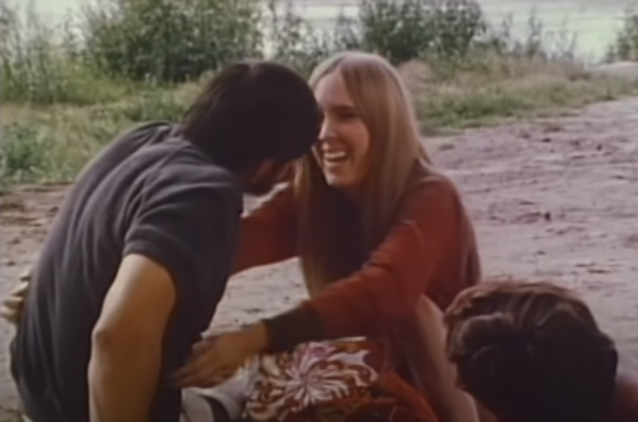 Jerry, with dark brown hair and friendly mutton chops, in a blue shirt, embracing Diane, with long blonde hair and a red shirt, also at the grassy picnic.