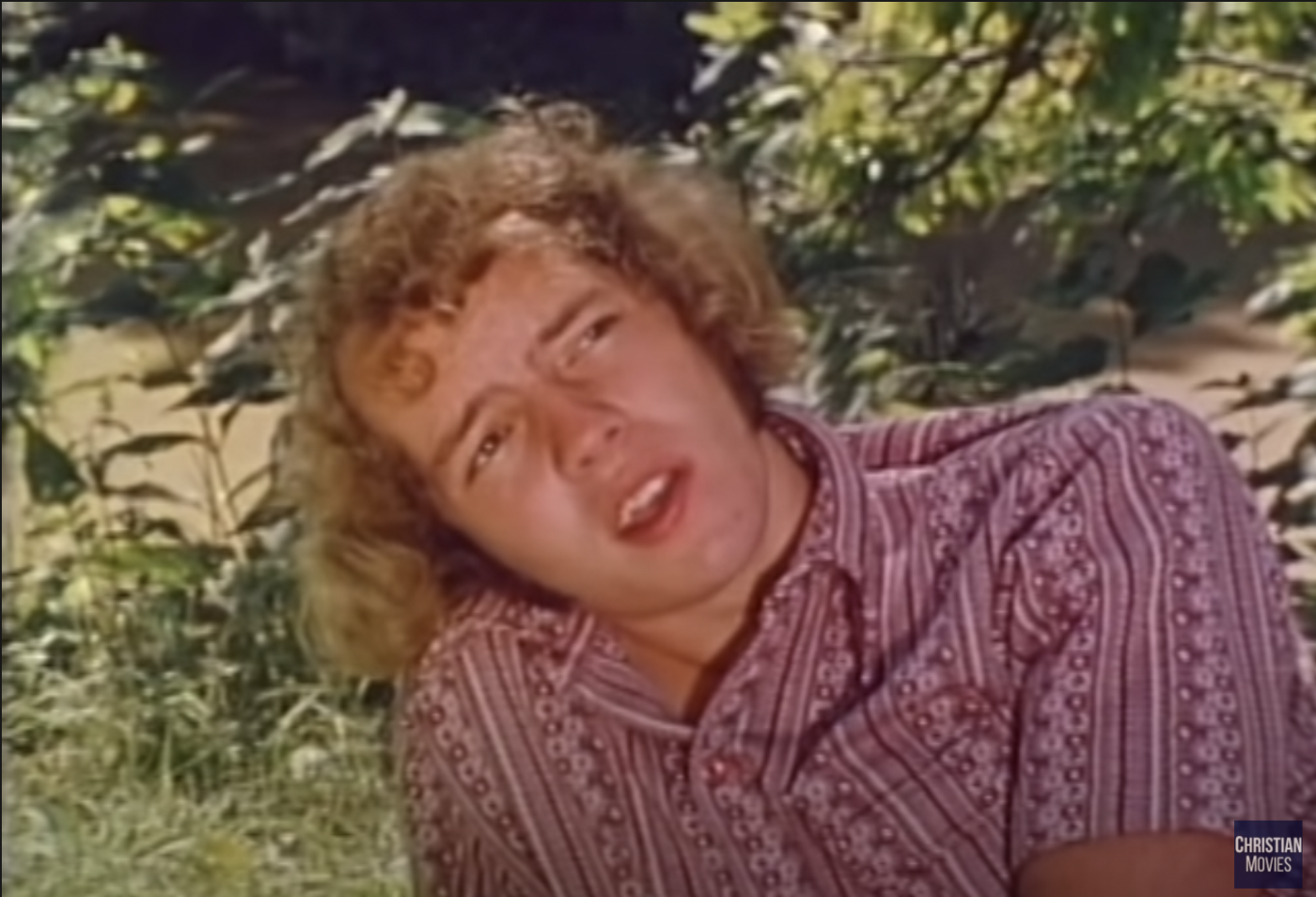 Duane, a young man with curly blond hair, looking quizzical and insufferable at the same time, with his mouth slightly open, in a grassy picnic setting, and wearing a patterned pink shirt.