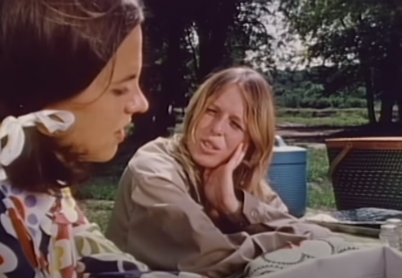 Two young women, Jenny, with brown hair, in the foreground and Patty, with blonde hair, in the background. Both are sitting at a picnic table. Jenny is wearing a shirt in a wild primary colour pattern I can't adequately describe and has a white ribbon in her hair. Patty is wearing a khaki shirt and leaning on the table and holding her face up with her hand.