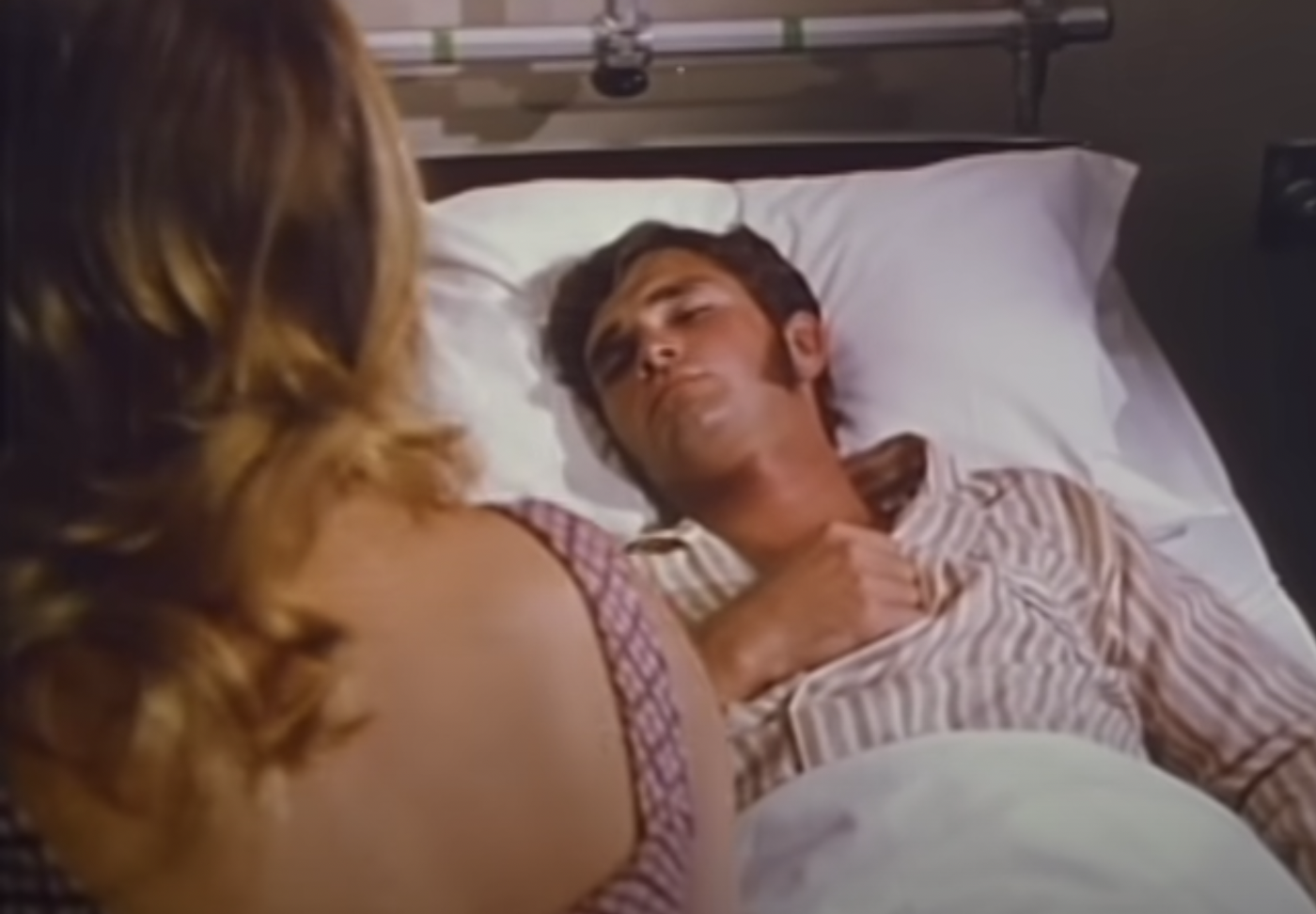 The back of Patty's head as Jim, who has brown hair and triangular sideburns, puts his fist on his chest in a hospital bed.