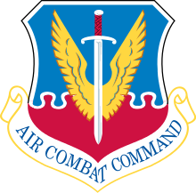 The exact same shape of shield as in the Trib Force crest, with a blue top half and a red bottom half, interlocking as if with jigsaw puzzles, and in front of it, a winged sword. The white scroll below this image reads "AIR COMBAT COMMAND," with the A and T in "COMBAT" even more disfigured by bending along with the scroll than in the other crest.