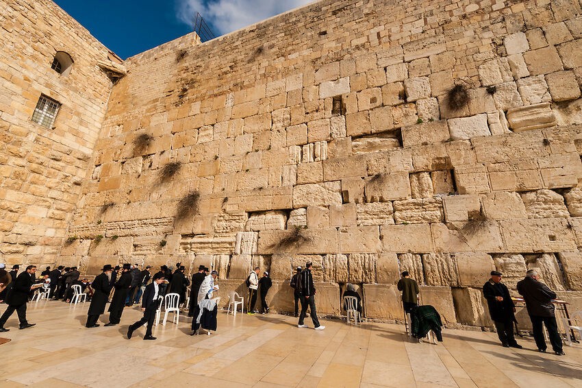 A bunch of people, mostly in black, many Orthodox Jews, in front of a corner of the Wailing Wall, which is made up of many different kinds of sandy brown rectangular stones, on a sunny day.