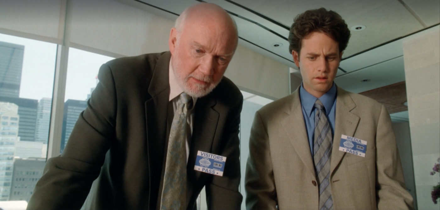 Chaim and Buck in an office, wearing suits with a visitor pass and a media pass pinned to their left breasts, respectively, looking down towards the camera, with windows showing office buildings in the background.