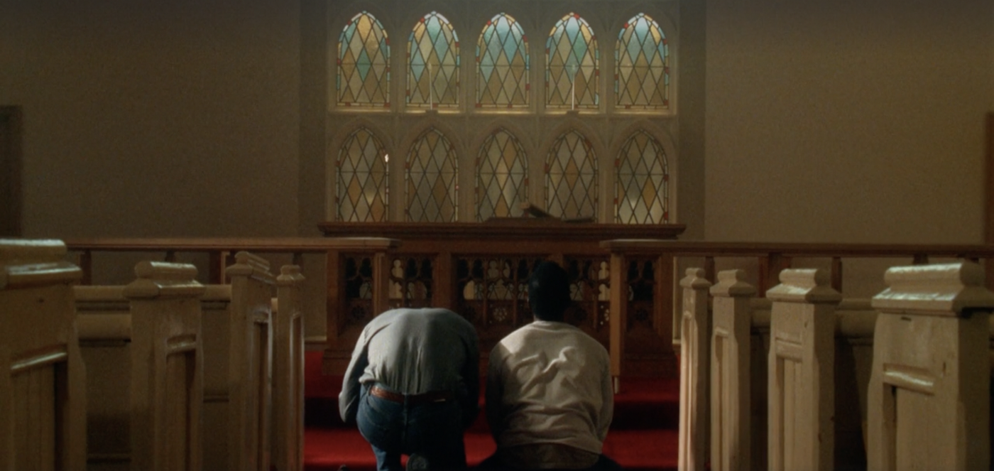 Rayford and Barnes kneeling, facing away from the camera and towards the altar, in a church.
