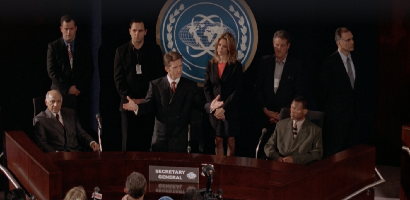 Nicolae gesturing to a room full of people, with a sign saying "Secretary General" in front of him, and a variation on the UN logo with a globe projection that is squashed vertically and flat ovoid rings encircling it, each on a diagonal so that they cross above and below the globe.