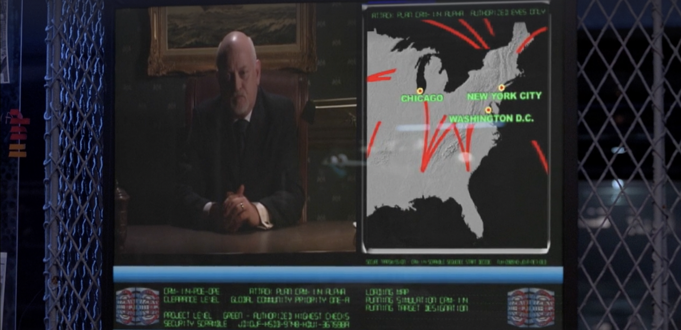On a video screen mounted to chain link fencing: a bald man in his sixties addressing the camera, some illegible green text on black, and a map showing red paths originating from within the US and heading towards Chicago, Washington, D.C., and New York City.