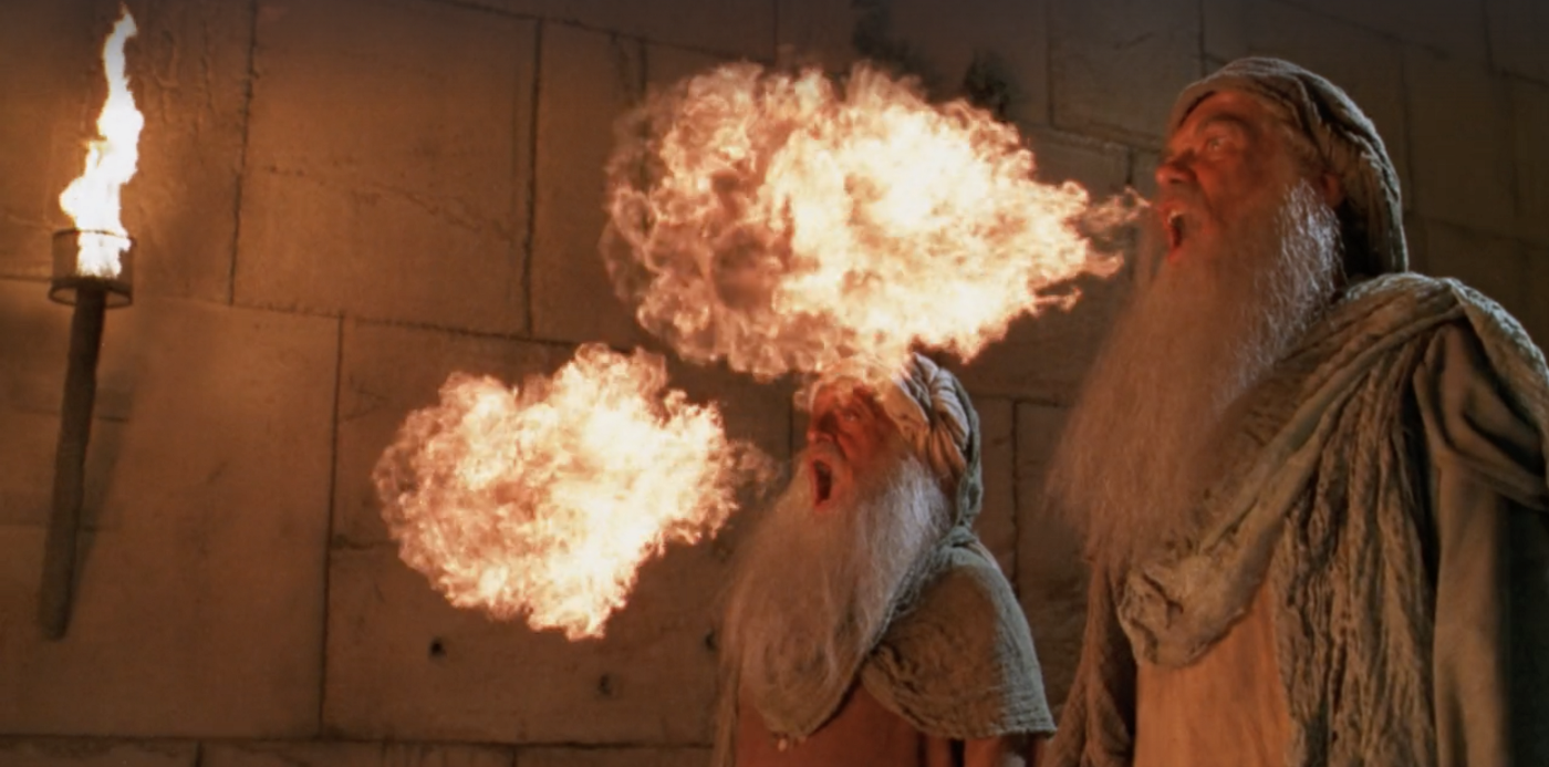 Two men dressed in rags breathing fire in front of a wall made of sand brown rectangular stones.