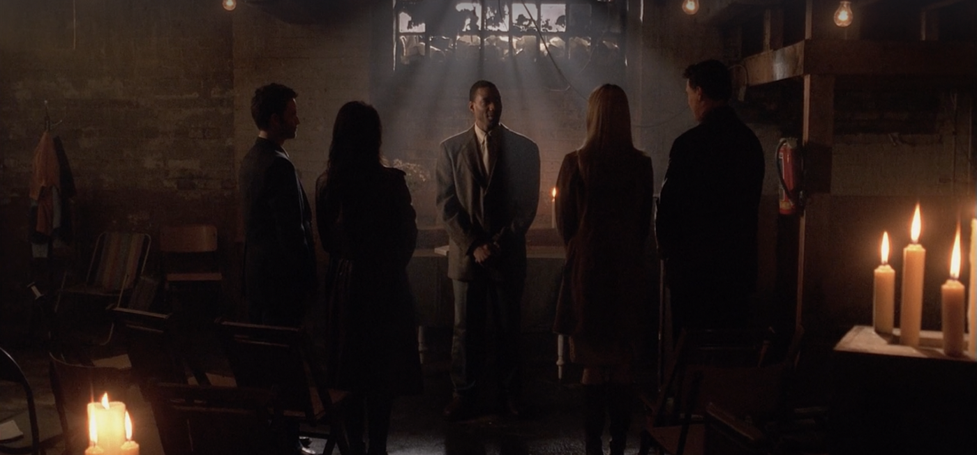 Pastor Barnes, now played by a different actor, addressing the two couples in the darkened church as light streams in from the window and candles light the foreground.