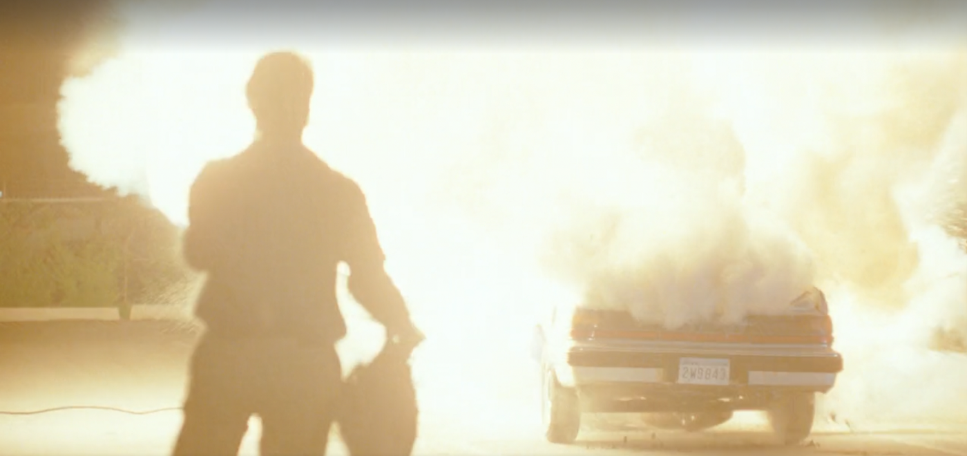 The silhouette of a figure holding a bag and looking at a car on fire.