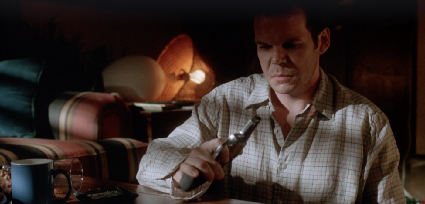 Chris inspecting a gun with a cup of coffee in front of him, with the light in the scene coming from a knocked over lamp in the back left.