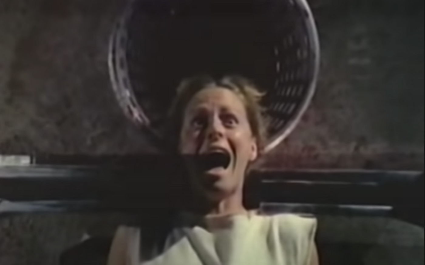 A guillotine's eye view of Patty screaming, face-up in the guillotine with her head over a bucket.