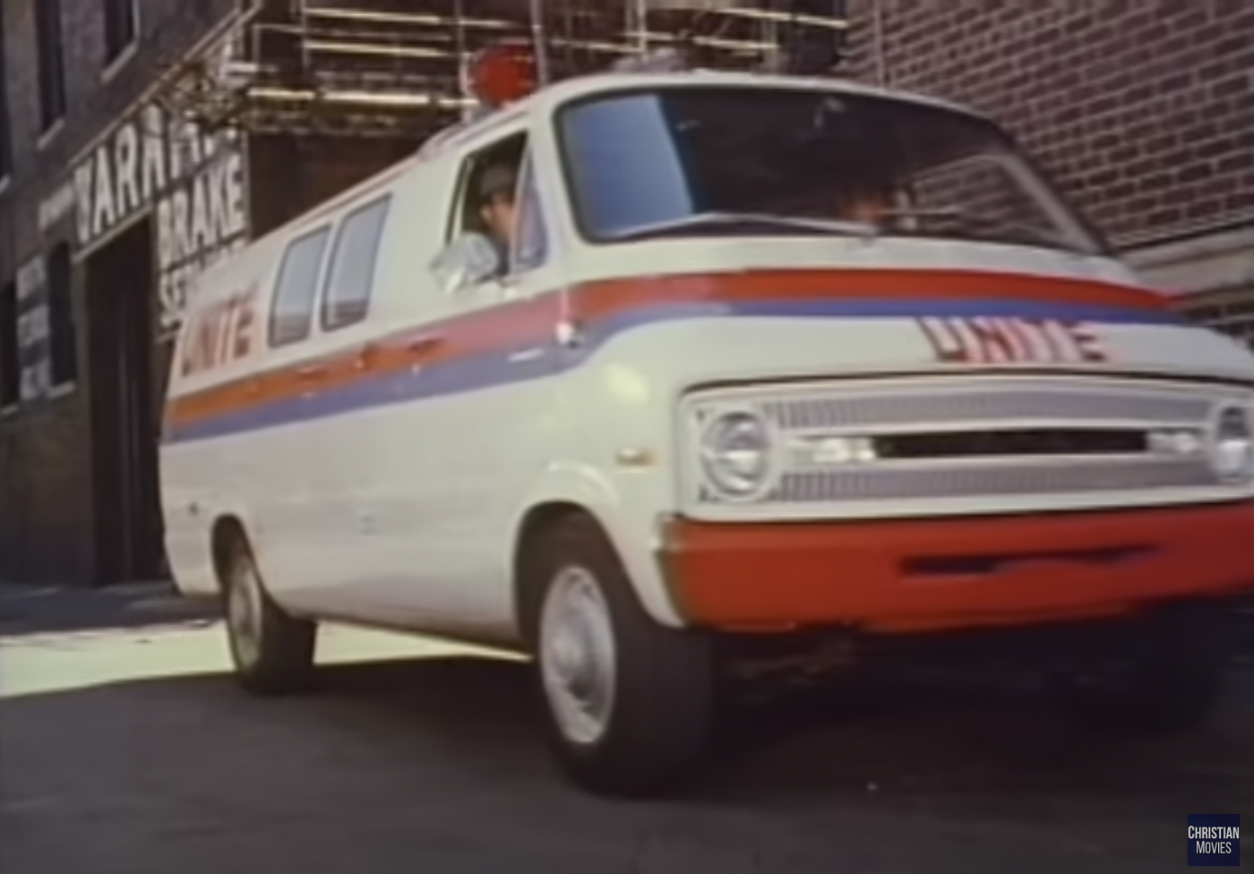 A white 1970s Dodge van with red and blue stripes along the side, a red bumper, and "Unite" in terrible lettering that looks like it's been made from red tape.