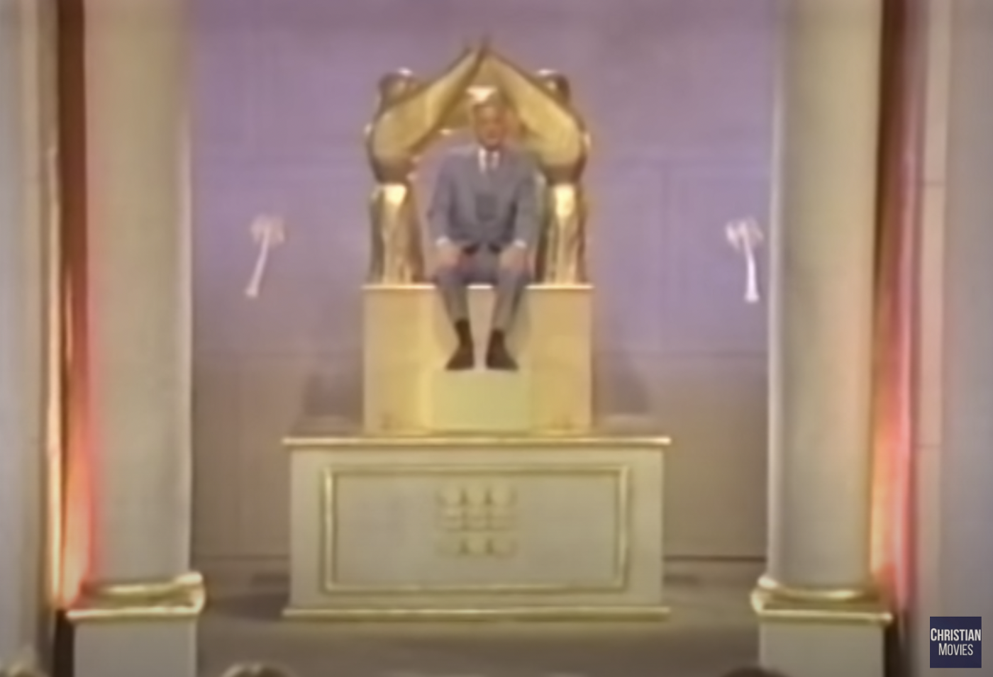 A blond man in a grey suit on a golden throne in a room with two palm trees painted on the  walls.