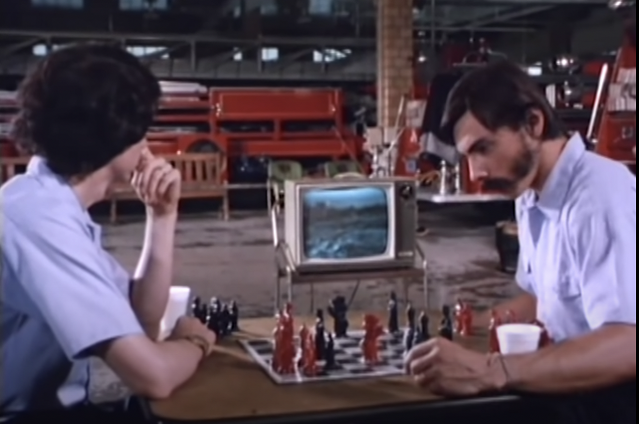 Jerry and another man playing chess with red and black pieces, wearing blue work uniform shirts.