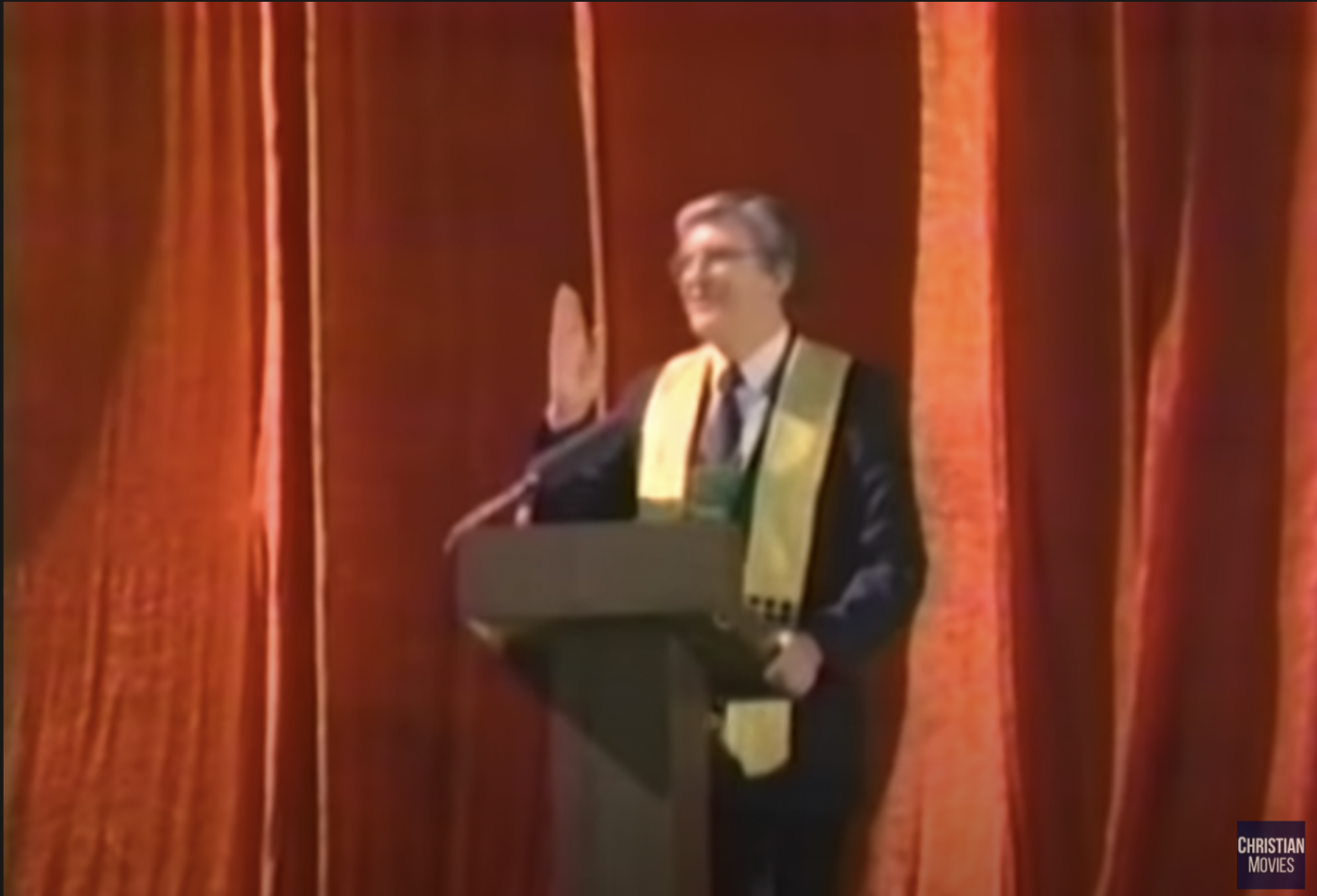 A grey-haired man holding his hand up in front of a lectern. He is wearing a sort of yellow scarf over his dark suit and there are orange-red curtains in the background. 