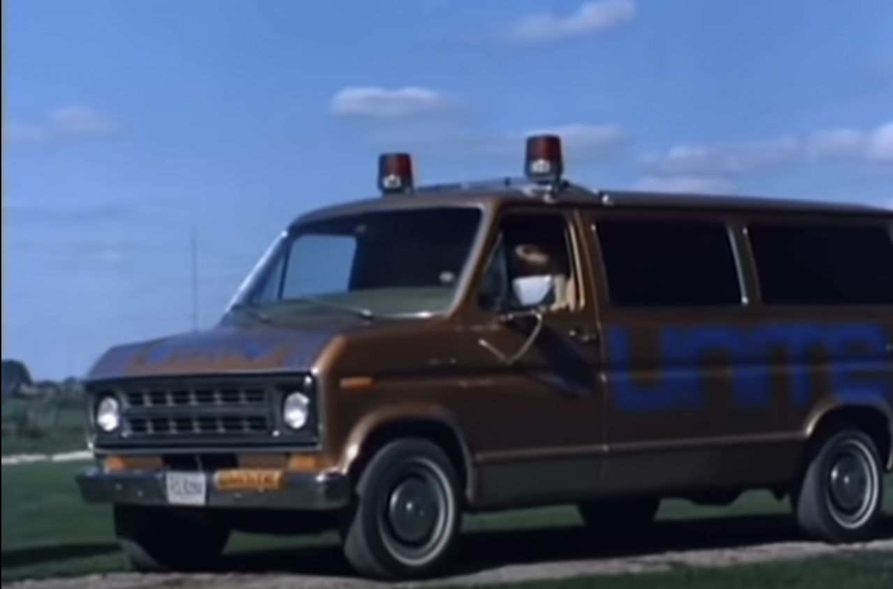 Another 1970s van. This one is brown and has heavy rust. UNITE is written in blue tape, even worse than last time. There are two police lights on the roof.