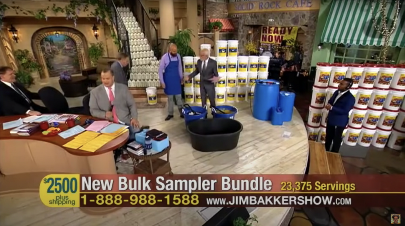 Jim Bakker and some other men in suits on the TV stage set of a living room that is filled with stacked buckets, gesturing at a big black mixing tub. The text at the bottom reads "$2500 plus shipping. New Bulk Sampler Bundle: 23,375 Servings. 1-888-988-15888 www.JIMBAKKERSHOW.com"