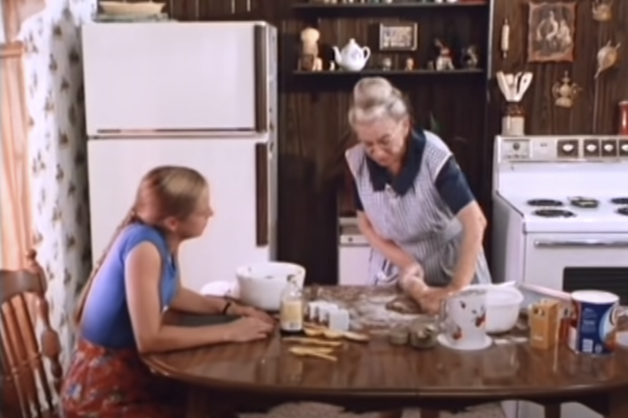 Patty sitting and listening to her grandmother as her grandmother, wearing an apron, rolls dough in a room with wood walls and lots of knick-knacks mounted on them.
