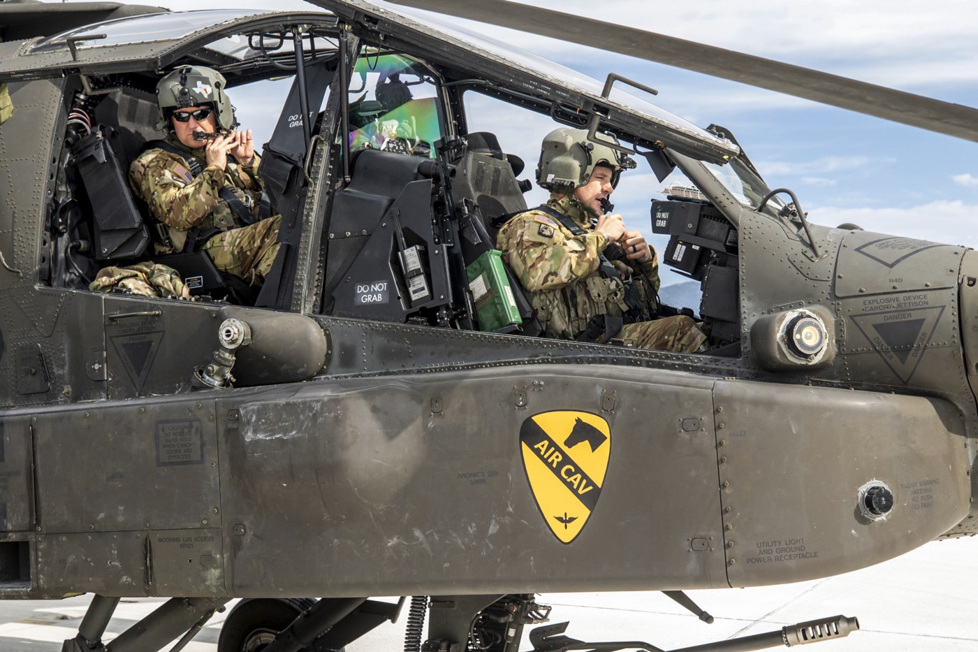 The pilot and weapons operator in the open cockpit of an AH-64 Apache helicopter, with a yellow "Air Cav" crest on the side featuring a horse head and a propellor with angelic wings. 