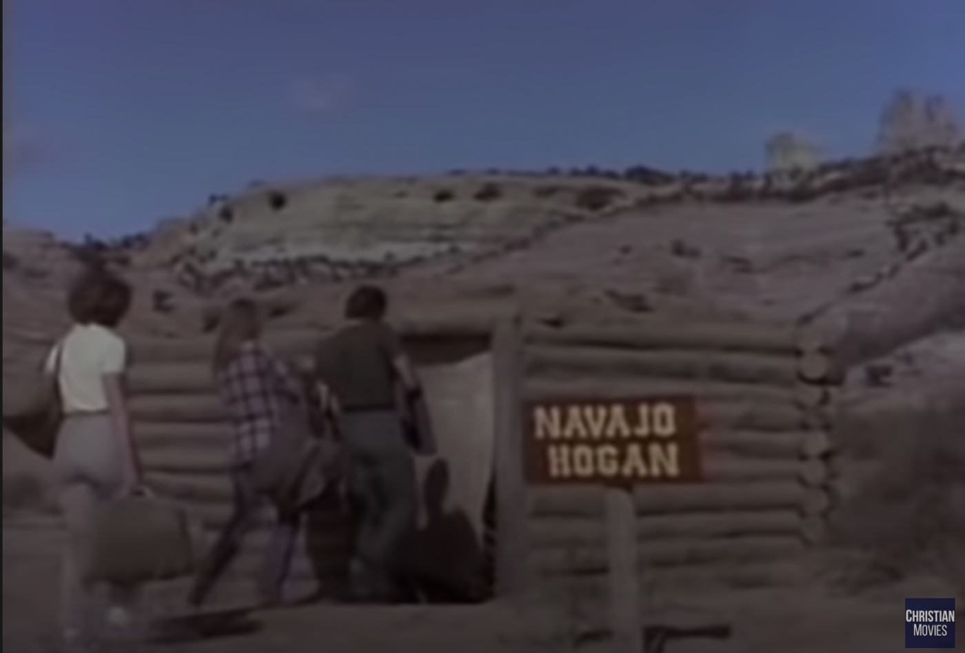 The crew entering a log dwelling with a sign in front of it that reads "Navajo hogan."