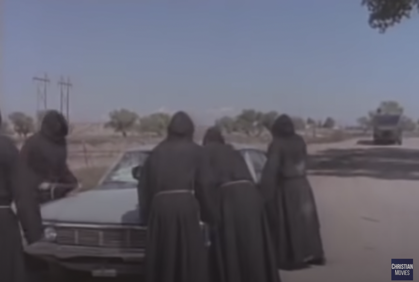 Five robed figures menacing a grey car with a smashed windshield on a dusty street as the RV approaches.