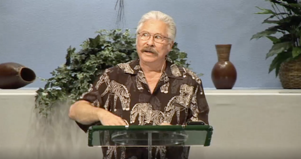 Hal Lindsey in the present day, still with a moustache and full head of hair, wearing a brown shirt with huge zebras on it, standing at a lectern, looking furious, with a knocked over plant in the background.