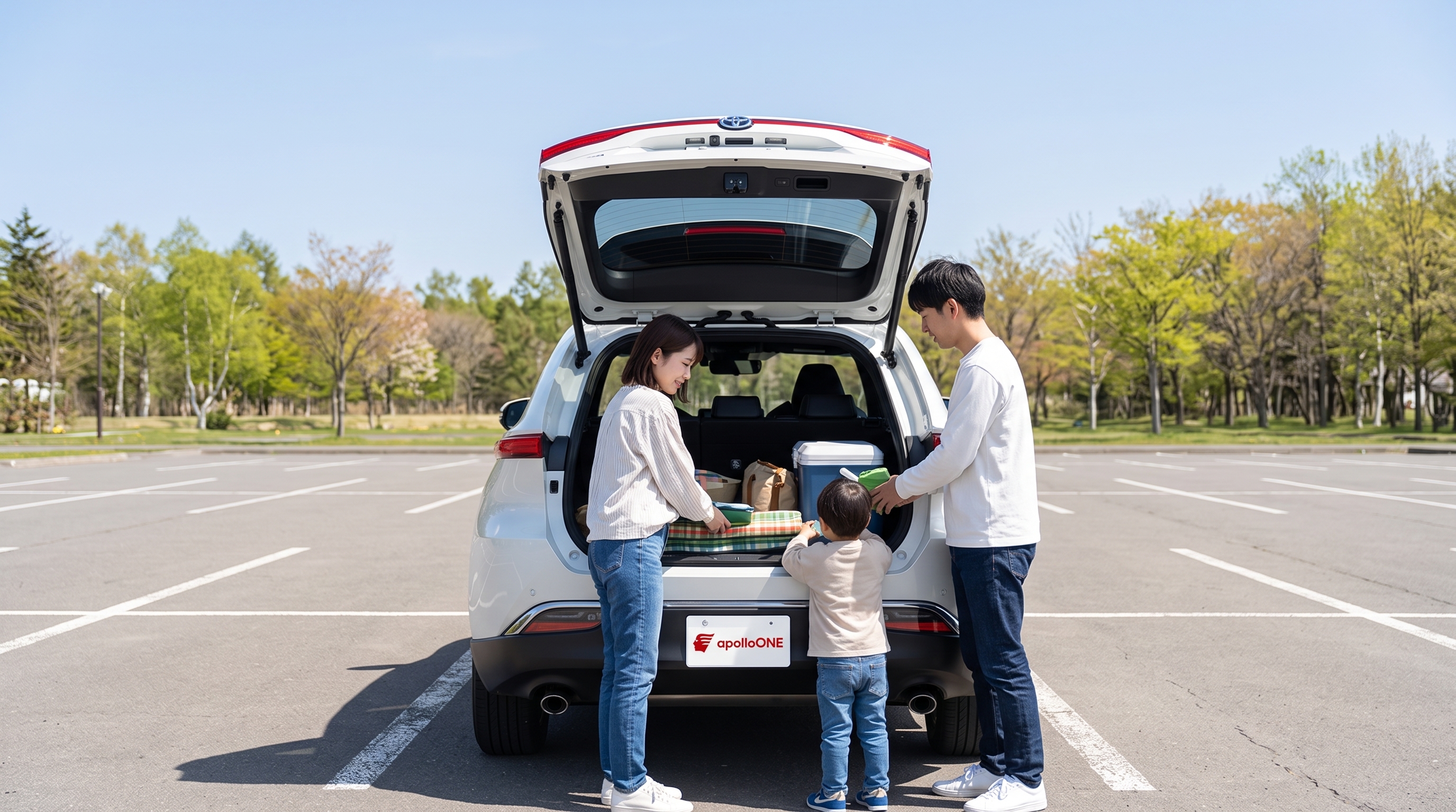 澄んだ青空の下の駐車場で、若い夫婦と小さな子供がSUVの大きく開いたバックドアにピクニック用の荷物を積み込んでいる、家族の日常的なお出かけを切り取ったシーン。