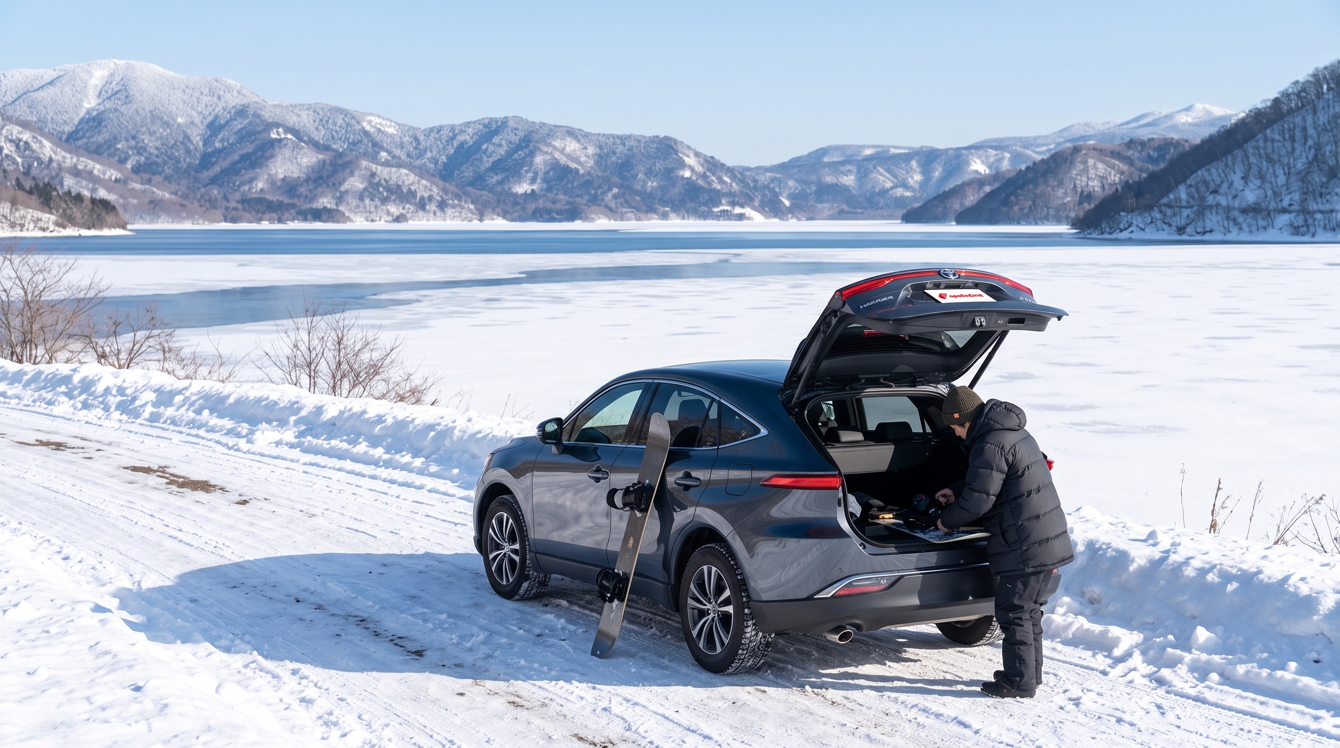 雪化粧をした冬の湖畔で、雪道に停まるグレーのSUVのバックドアを開け、ダウンジャケットを着た人物がスノーボードの準備をしているアウトドアでの体験シーン。