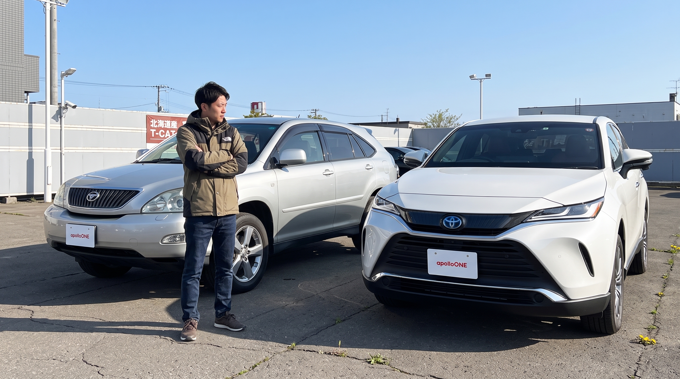 春の爽やかな青空の下、屋外スペースに並んで駐車された白いSUV車とシルバーのSUV車の前で、男性客が腕を組みながら2台のフォルムを見比べているシーン。