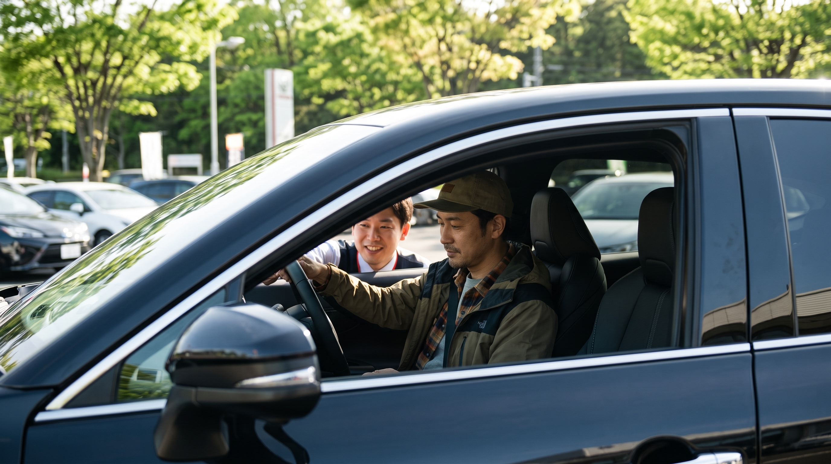 新緑の木漏れ日が輝く駐車場で、SUVに乗る男性客と窓越しに笑顔で話すスタッフの様子。試乗への期待感を高める、二人の自然なコミュニケーションを捉えたシーンです。