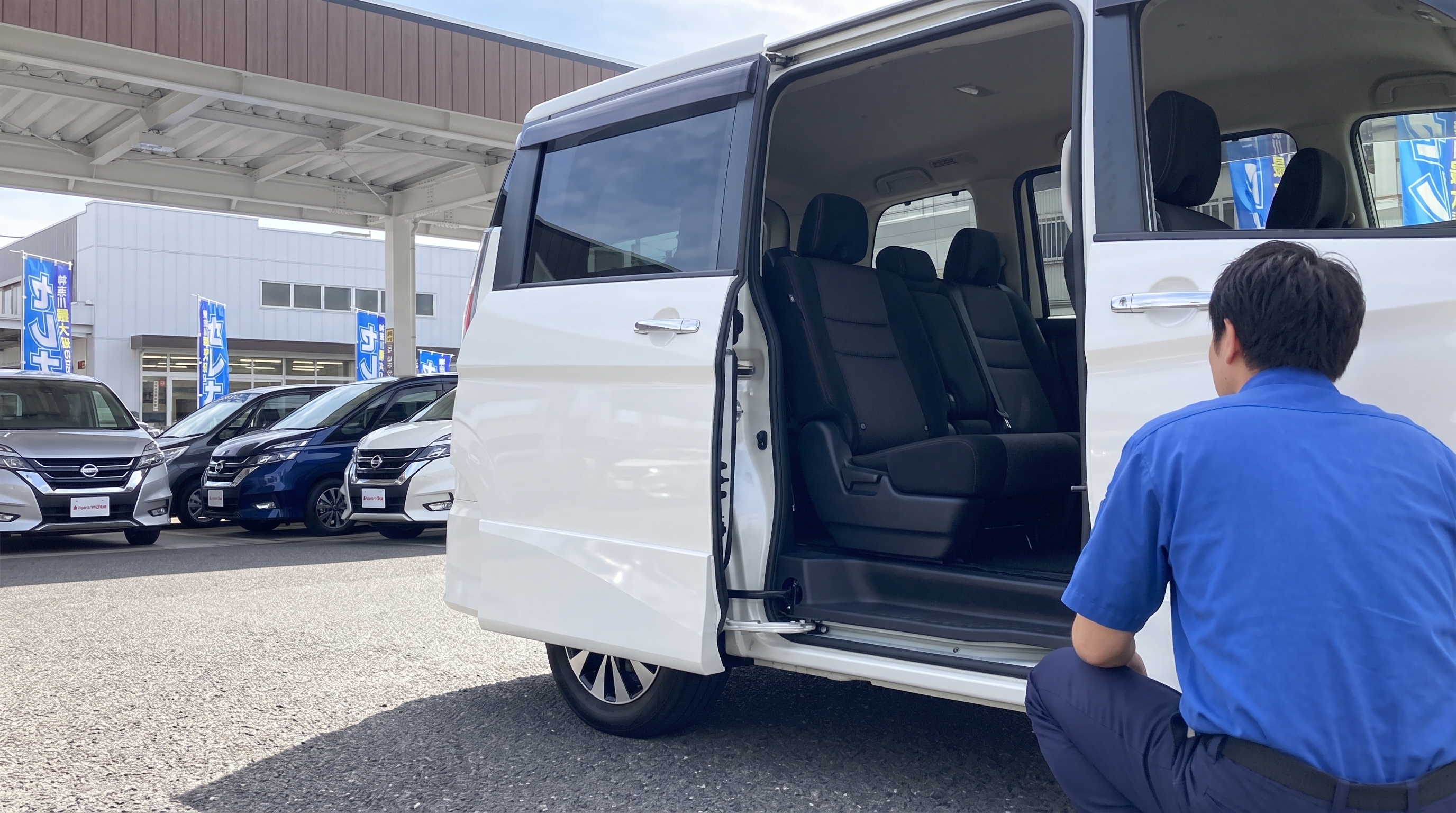 初夏の日差しが差し込む展示場で、スライドドアが開いたパールホワイトのミニバンの清潔な内装を、査定士がしゃがみ込んで下から丁寧にチェックしているシーン。