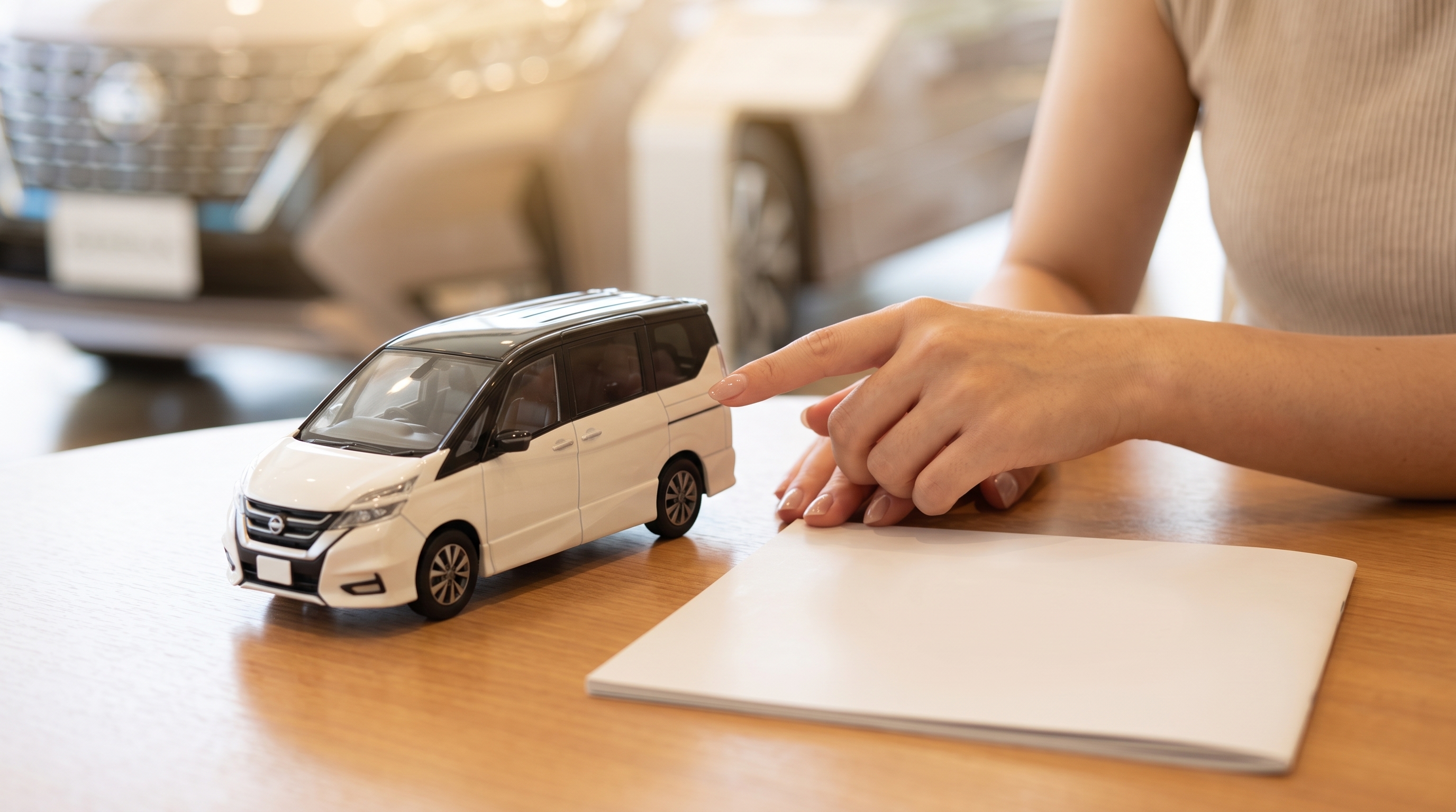 明るいショールームで、女性がテーブルに置かれた車の色見本模型を指差している手元のクローズアップ。傍らにはパンフレットがあり、背景には展示車がボケて写っている。