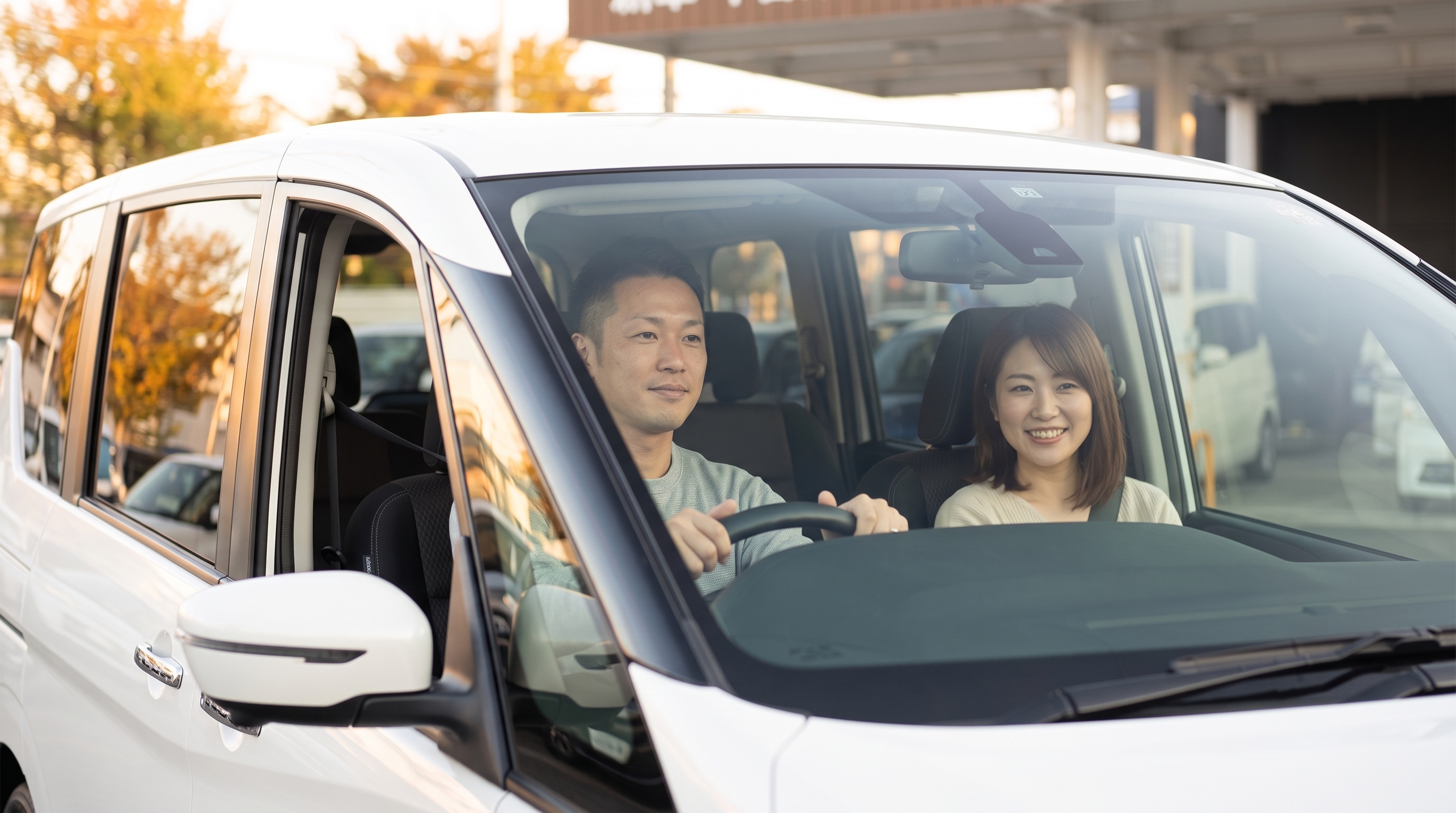 秋の夕暮れ前、試乗に出発するミニバンの運転席に座る父親と助手席で微笑む母親を、車の外から窓越しに捉えた、広々とした車内の和やかな雰囲気が伝わるシーン。