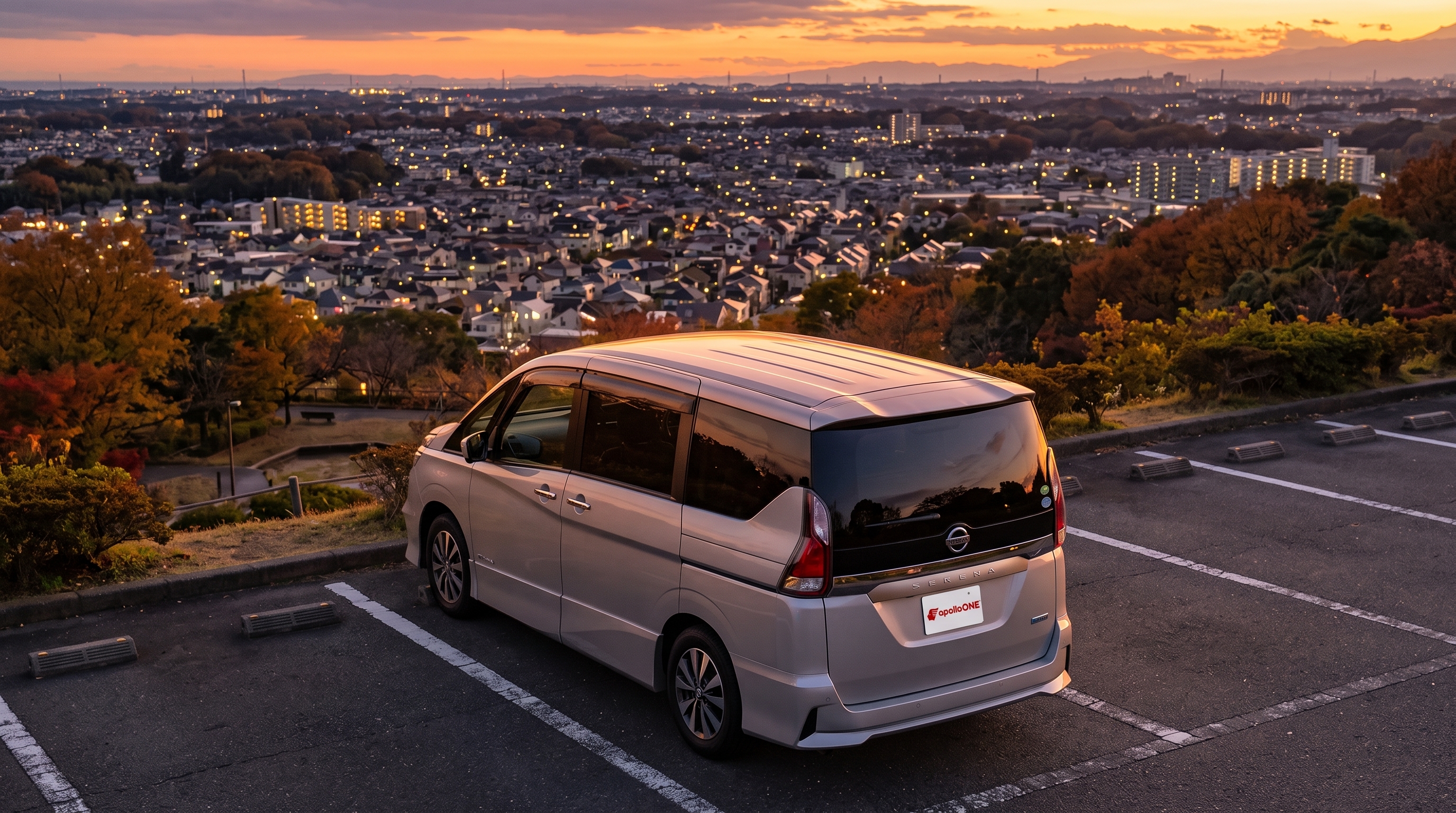 秋の夕暮れ時、見晴らしの良い高台の駐車場に停められたシルバーのミニバンの様子。夕日に染まる車体と奥に広がる住宅街の灯りを俯瞰で捉えた、暖かみのある日常風景。