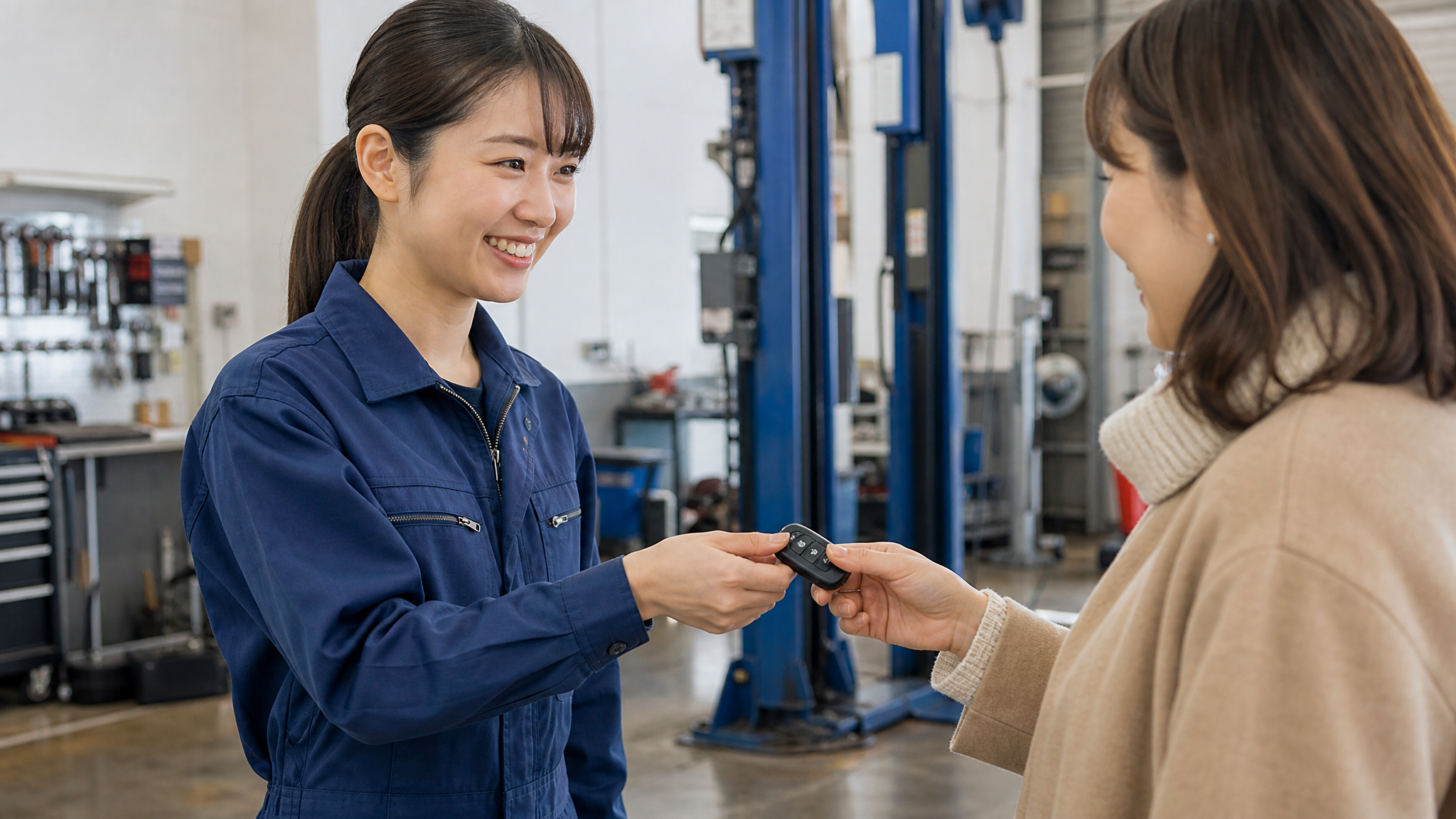 冬の午後の穏やかな光の中、女性が整備士からスマートキーを受け取り、安心した笑顔を見せているシーン。背景には整備ピットの工具や整備用リフトが少し見えている様子。