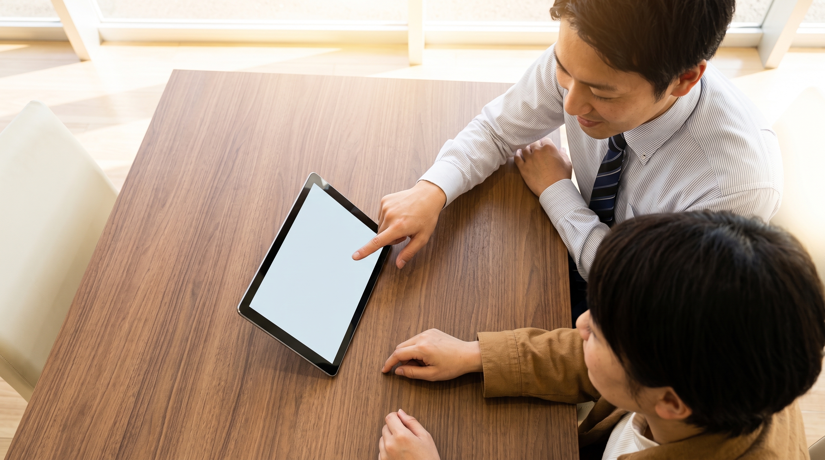 明るいショールームの商談スペースで、スタッフが木目調テーブル上のタブレットを指差して買取相場を説明し、顧客がそれを見つめる様子を真上から捉えたシーン。