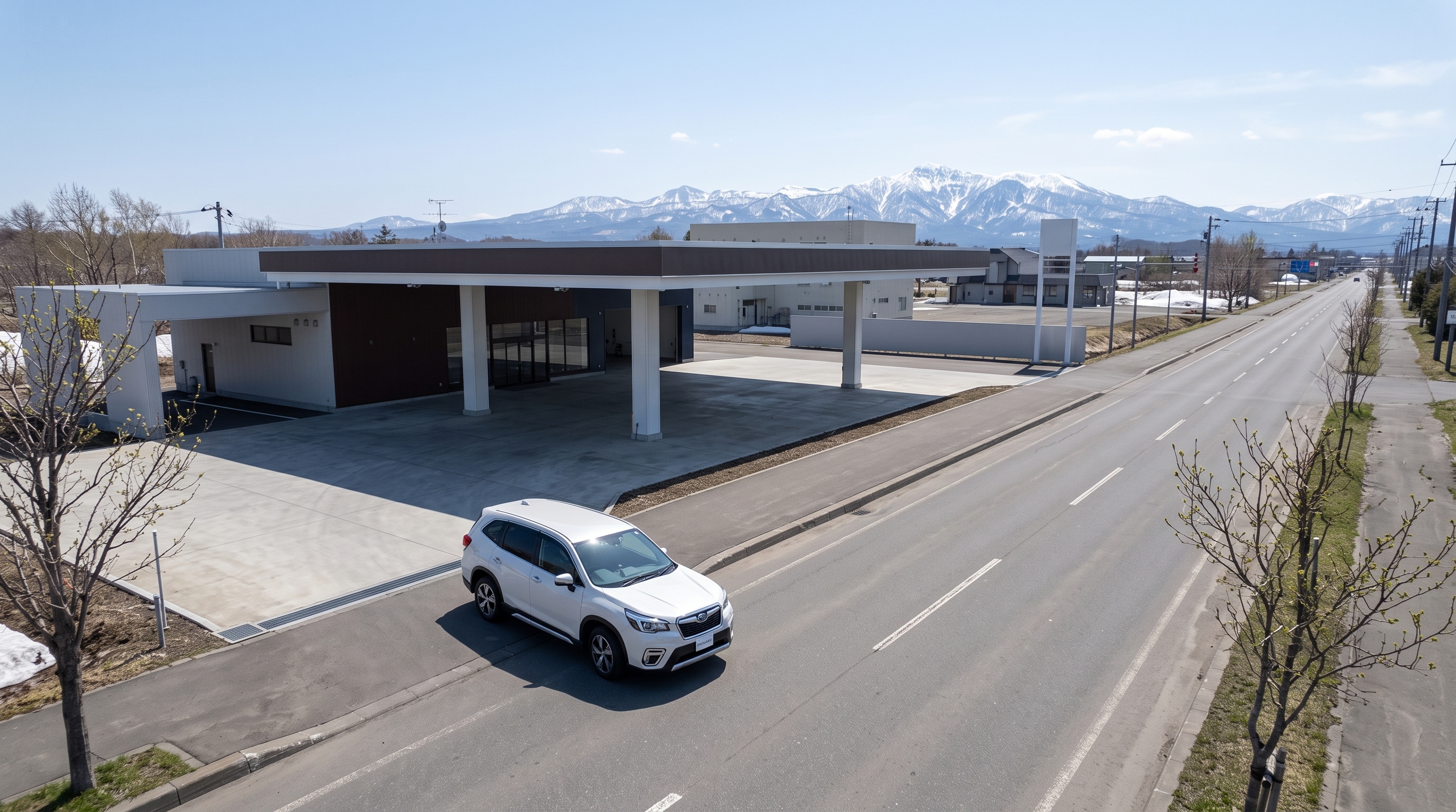 春の穏やかな昼下がり、遠くに山の稜線を望む広々とした道路へ向けて、乾いたアスファルトの上を颯爽と走り出す白いSUVを捉えた俯瞰構図の風景。