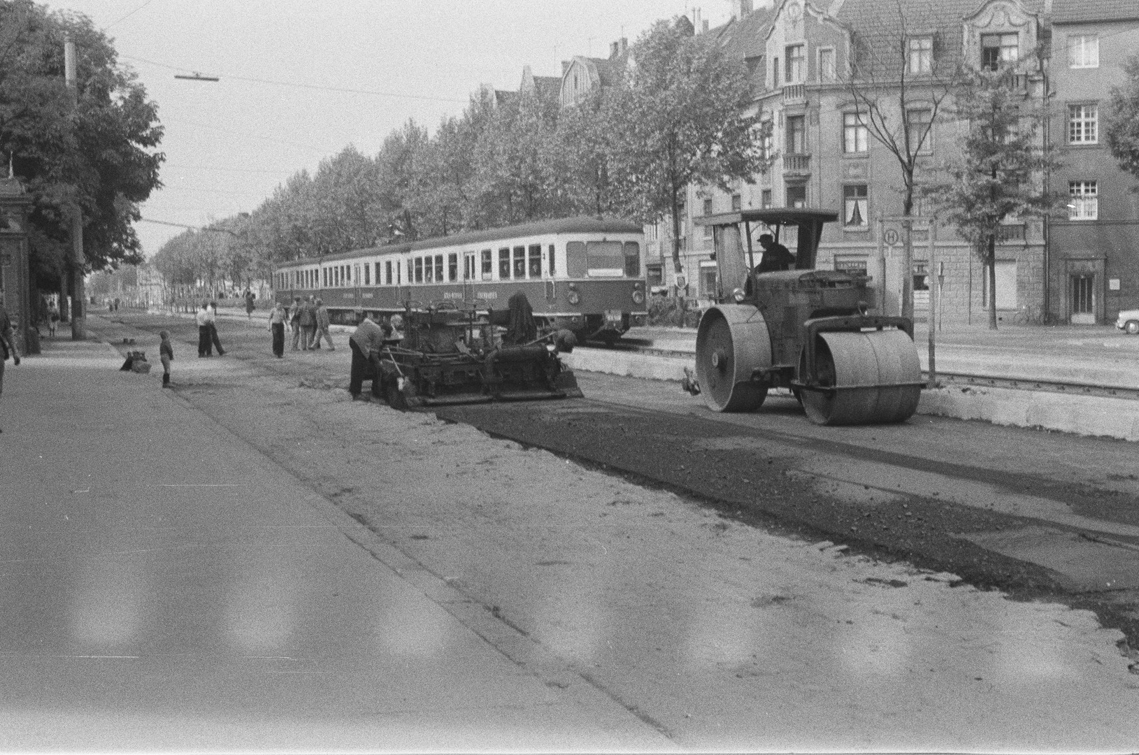 Straßenbauarbeiten: Die Luxemburger Straße wird asphaltiert (Richtung stadtauswärts fotografiert). Ganz links ist ein kleines Stück der Außenummauerung des Weißhaus-Schlosses zu sehen und auf den Gleisen die „Köln-Bonner Eisenbahn“ (KBE). Deren Schienen endeten damals am Barbarossa-Platz. Köln 1958 oder 1959 (Zimmermann hat eine 17 Fotos umfassende Dokumentation dieser Straßenbauarbeiten aufgenommen, die im weiteren Verlauf dieser allmählich wachsenden digitalen Ausstellung vollständig gezeigt werden soll.)