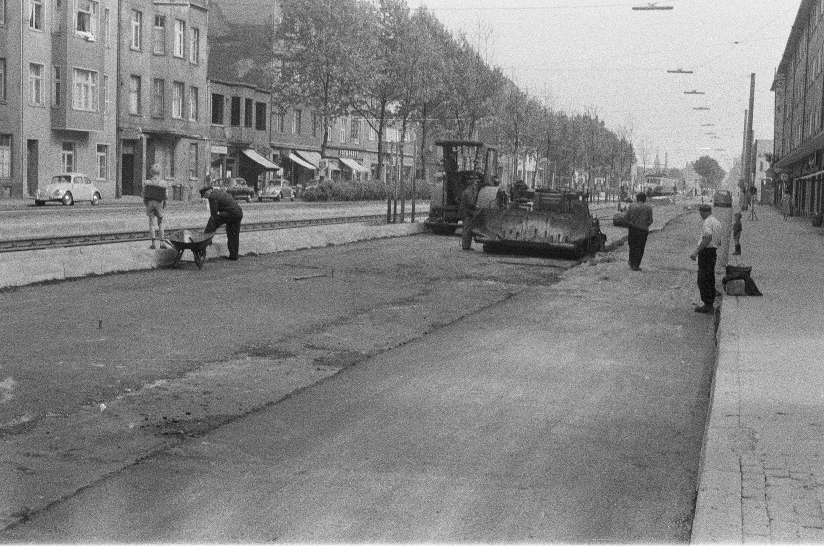 Straßenbauarbeiten: Die Luxemburger Straße wird asphaltiert (Richtung stadteinwärts fotografiert). Der Junge mit Schulranzen blickt auf die Häuserzeile, in der deutlich Kriegsschäden zu sehen sind: Die oberen Stockwerke eines offensichtlich nicht mehr bewohnbaren Hauses fehlen, im Erdgeschoss scheint jedoch ein Geschäft in Betrieb zu sein. Im Hintergrund rechts die KBE. Köln 1958 oder 1959